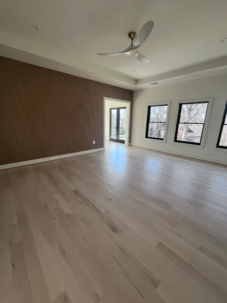 A spacious, empty living area featuring freshly swept light wood floors, bright black-framed windows, and a dark accent wall after a professional cleaning by L&V Cleaning.