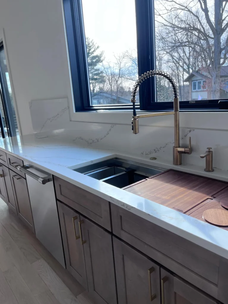 A pristine kitchen sink area featuring wiped-down white quartz countertops, a brass faucet, and clean black-framed windows, demonstrating the deep cleaning results of L&V Cleaning.
