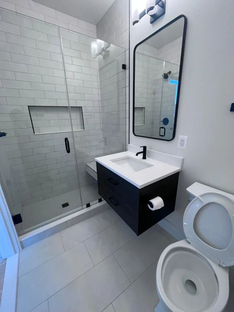 A compact modern bathroom featuring a streak-free glass shower enclosure, a spotless black floating vanity, and shining white floor tiles after a professional cleaning by L&V Cleaning.
