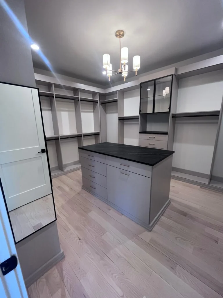 A large, luxury walk-in closet featuring pristine custom grey shelving, a center island with a dark stone countertop, and spotless light wood floors after a detailed clean by L&V Cleaning.