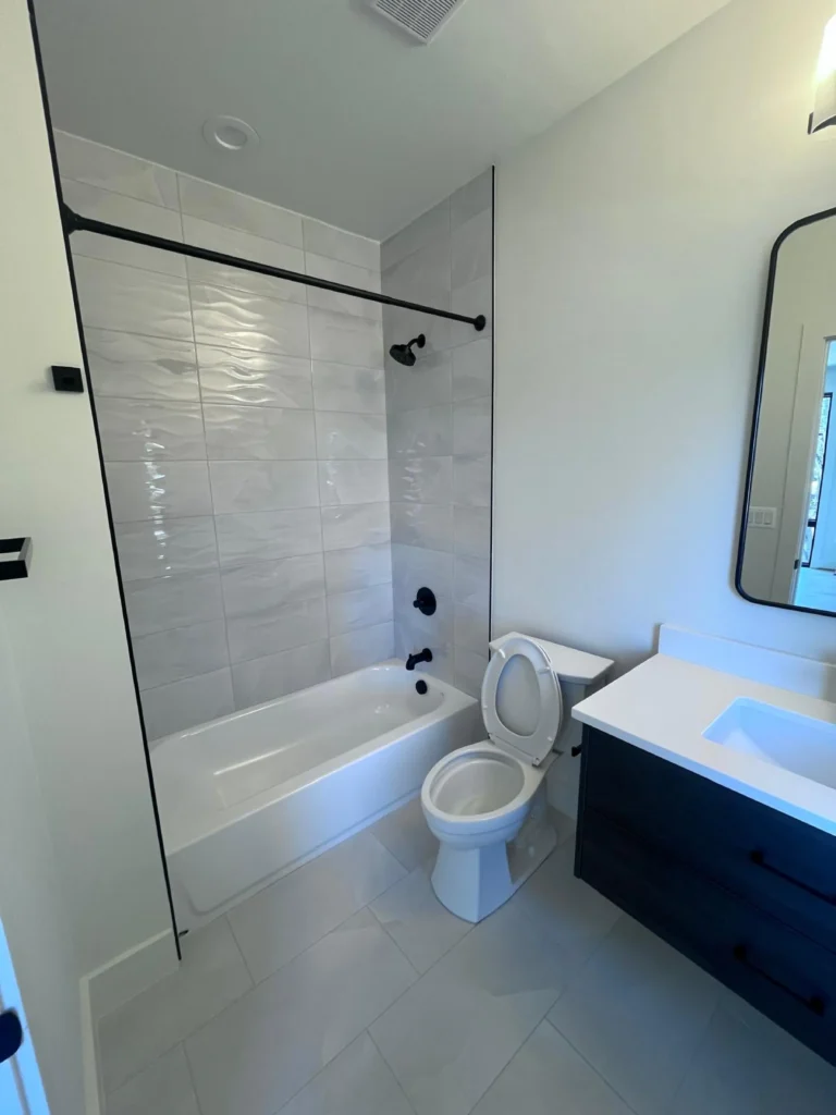 A bright, spotless bathroom featuring a white bathtub, grey textured shower tiles, matte black hardware, and a clean dark vanity, maintained by L&V Cleaning.