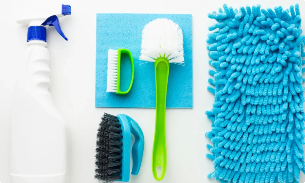 An flat lay arrangement of essential home cleaning supplies including a white spray bottle, blue microfiber cloth, green and black scrubbing brushes, and a fluffy blue duster, matching the recommended tools for Northern Virginia homeowners.