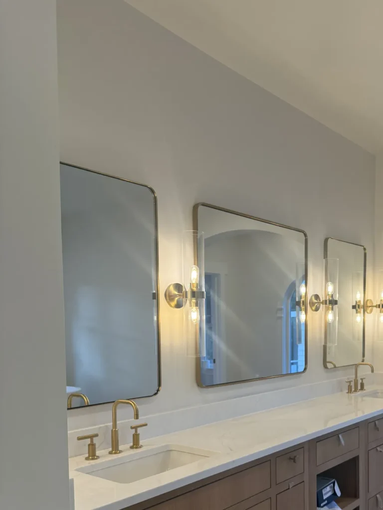 A modern bathroom vanity featuring a spotless white countertop, polished brass faucets, and streak-free mirrors after a deep clean by L&V Cleaning.