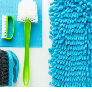 An flat lay arrangement of essential home cleaning supplies including a white spray bottle, blue microfiber cloth, green and black scrubbing brushes, and a fluffy blue duster, matching the recommended tools for Northern Virginia homeowners.