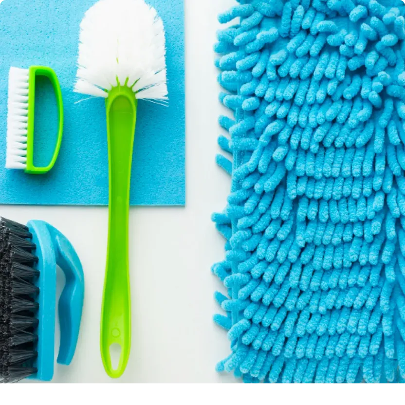 An flat lay arrangement of essential home cleaning supplies including a white spray bottle, blue microfiber cloth, green and black scrubbing brushes, and a fluffy blue duster, matching the recommended tools for Northern Virginia homeowners.