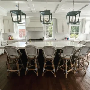 A bright, meticulously clean luxury kitchen in a Northern Virginia home featuring white cabinets, a large center island with five wicker barstools, dark hardwood floors, and modern hanging light fixtures, demonstrating residential deep cleaning results by L&V Cleaning.