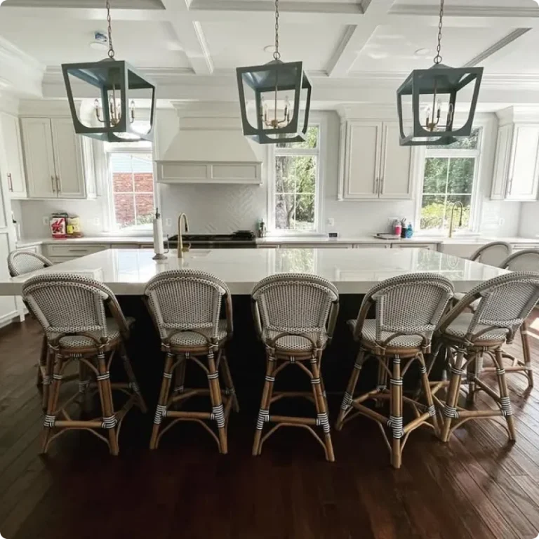 A bright, meticulously clean luxury kitchen in a Northern Virginia home featuring white cabinets, a large center island with five wicker barstools, dark hardwood floors, and modern hanging light fixtures, demonstrating residential deep cleaning results by L&V Cleaning.