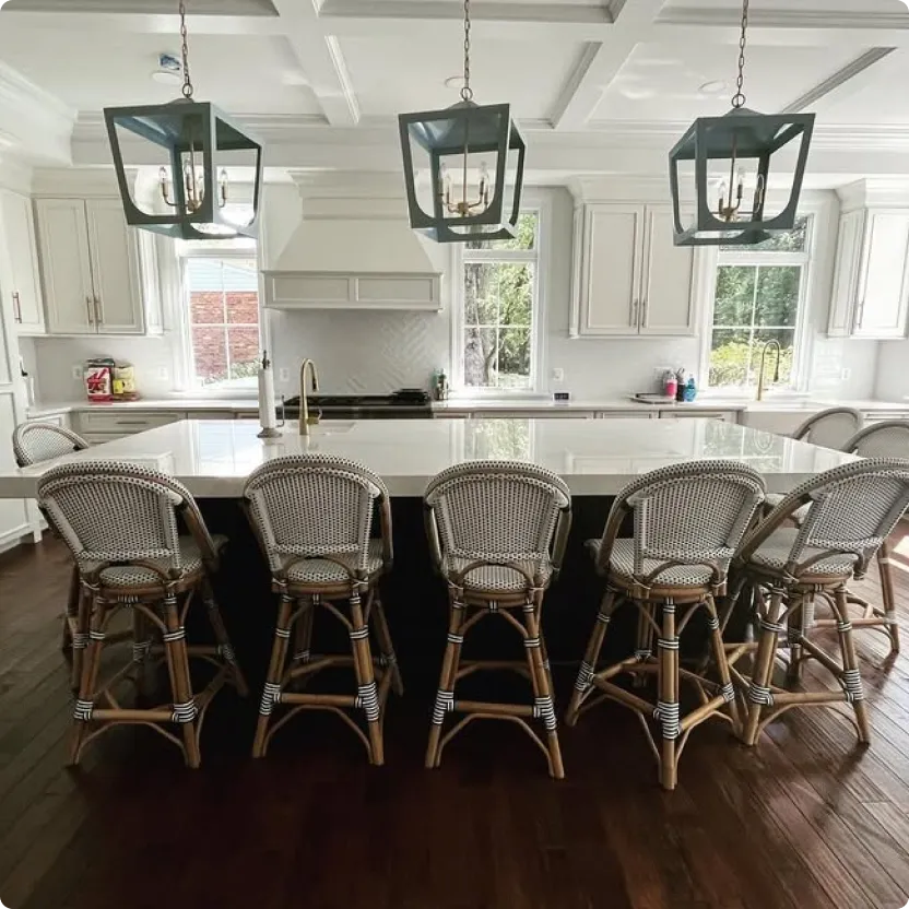 A bright, meticulously clean luxury kitchen in a Northern Virginia home featuring white cabinets, a large center island with five wicker barstools, dark hardwood floors, and modern hanging light fixtures, demonstrating residential deep cleaning results by L&V Cleaning.