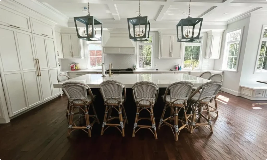 A bright, meticulously clean luxury kitchen in a Northern Virginia home featuring white cabinets, a large center island with five wicker barstools, dark hardwood floors, and modern hanging light fixtures, demonstrating residential deep cleaning results by L&V Cleaning.